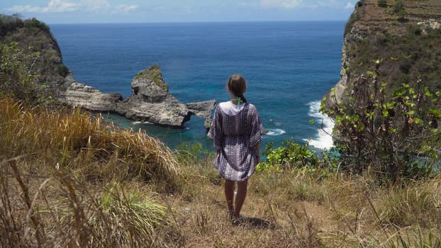 Young girl stands on the edge of a cliff and looks at the sea. Girl on the edge of the cliff enjoys the view of the ocean. Atuh beach on Nusa Penida island. Travel concept.