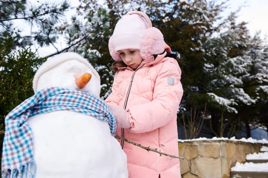 Charming cute little child girl puts on a blue shawl on a snowman, during active winter leisure games in the snowy backyard in the fresh air. Happy childhood and healthy lifestyle. Christmas time