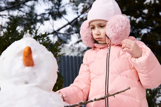 Adorable lovely kid girl in pink down jacket and fluffy earmuffs, building snowman, enjoying happy winter holidays outdoors. Childree. Happy carefree childhood. Recreation. Lifestyle. Leisure activity