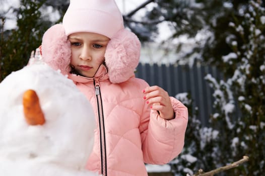 Caucasian adorable child 6 years old, lovely little girl in pink warm winter clothes and fluffy ear muffs, building a snowman in the snow covered backyard. Winter leisures activity. Christmas time