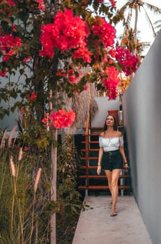 A peaceful woman in a white top and black shorts walks along a path with pink flowers in full bloom, basking in the radiant sunlight, while surrounded by a picturesque landscape of cascading blossoms