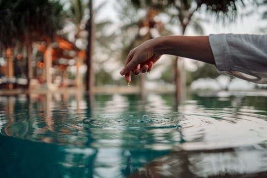 An elegant woman's hand touches the surface of the turquoise water, creating ripples, symbolizing the connection between humans and nature.