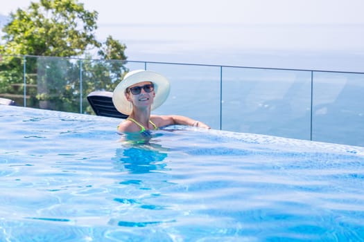 Summer travel vacation concept, Happy traveler asian woman with hat and bikini relax in luxury infinity pool