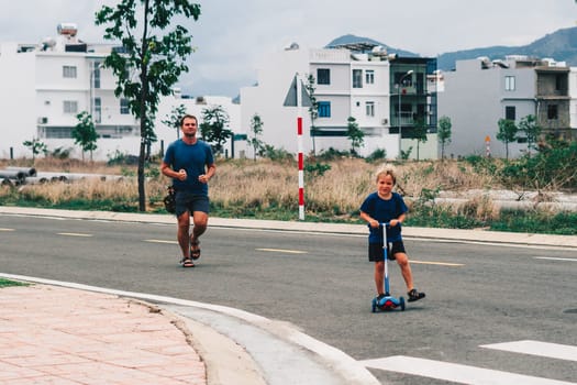 Father son go for sports, morning jog, man runs, child rides a scooter, modern village.