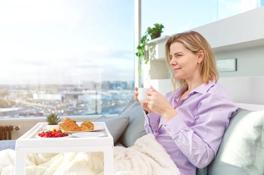 Happy middle aged, beautiful woman having breakfast, relaxing in hotel bed. Woman having breakfast sitting on her bed in luxury apartments. enjoying lazy morning lifestyle