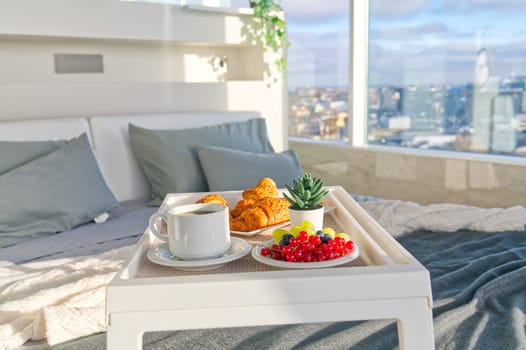 White Tray with tasty breakfast on bed in light bedroom with big windows in skyscraper. white table with tasty breakfast in bed at bed room in skyscraper