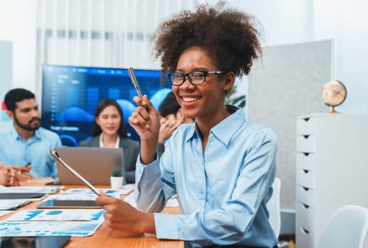 Happy young african businesswoman wearing glasses portrait with group of office worker on meeting with screen display business dashboard in background. Confident office lady at team meeting. Concord