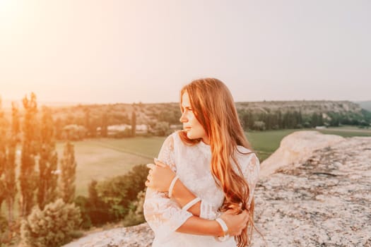 Romantic beautiful bride in white dress posing with sea and mountains in background. Stylish bride standing back on beautiful landscape of sea and mountains on sunset