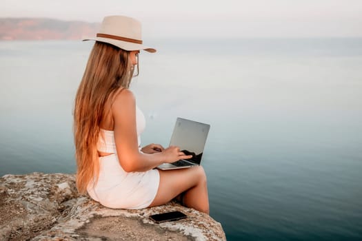 Digital nomad, Business woman working on laptop by the sea. Pretty lady typing on computer by the sea at sunset, makes a business transaction online from a distance. Freelance, remote work on vacation