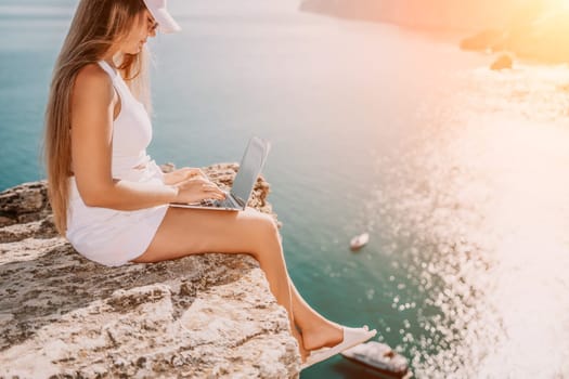 Successful business woman in yellow hat working on laptop by the sea. Pretty lady typing on computer at summer day outdoors. Freelance, travel and holidays concept.