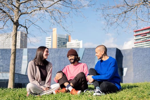 three young friends laughing happy sitting in the lawn at the city park in a sunny day, concept of friendship and urban lifestyle, copy space for text