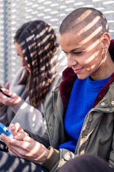 young woman with shaved head using mobile phone with friends sitting in the floor at city, concept of technology of communication and modern lifestyle, copy space for text