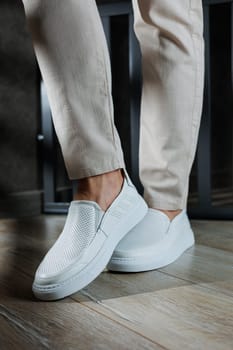 Close-up of male feet in white casual shoes. Fashionable young man standing in leather stylish white moccasins in fashionable trousers. Seasonal summer men's shoes. Casual street style.