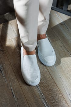 Close-up of male feet in white casual shoes. Fashionable young man standing in leather stylish white moccasins in fashionable trousers. Seasonal summer men's shoes. Casual street style.