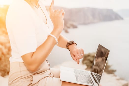 Successful business woman in yellow hat working on laptop by the sea. Pretty lady typing on computer at summer day outdoors. Freelance, travel and holidays concept.