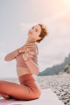 Young woman with long hair in white swimsuit and boho style braclets practicing outdoors on yoga mat by the sea on a sunset. Women's yoga fitness routine. Healthy lifestyle, harmony and meditation