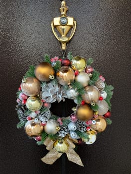 Luxurious Christmas decorative wreath, handmade with pine cones and golden balls, hangs on the dark front door of the house.