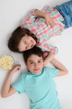 View from above of two adorable kids, mischievous little child girl sister and her older brother - preteen boy eating pop corn and smiling looking at camera, lying on white isolated studio background