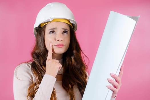 Thoughtful young girl architect in a hard hat holds her project in her hands on the pink background. Student project concept.