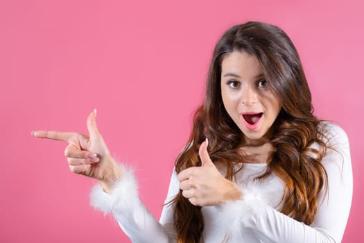 A portrait of a young lady looks at the camera and stands on a pink backdrop, gesturing towards copy space.