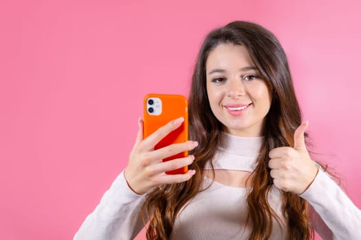 Young woman showing thumb up in like or approval of new good, holding a smartphone standing on the pink background.