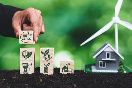 Businessman putting Net Zero symbol cube on stack top for corporate environmental protection and sustainability. Modern house model power by clean wind energy with net zero CO2 emission concept. Alter