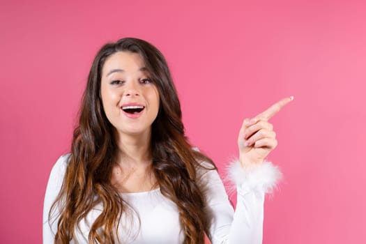 A young and joyful woman radiates a bright smile standing against a vibrant pink backdrop, extending her finger to indicate the copy space for advertising purposes.