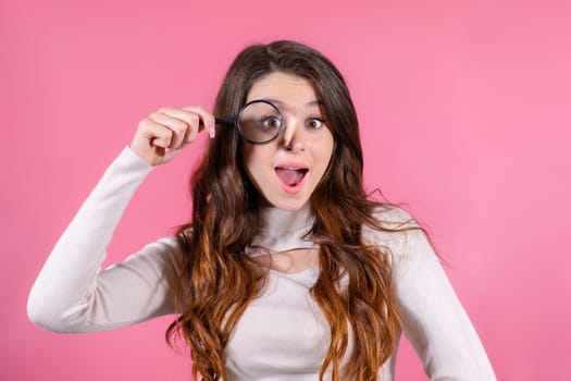 Funny woman looking through magnifying glass against pink background standing in white sweater. Searching information concept.