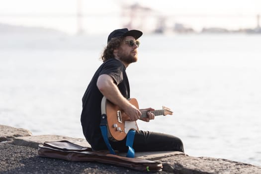 A man hipster playing electric guitar at the waterfront. Mid shot