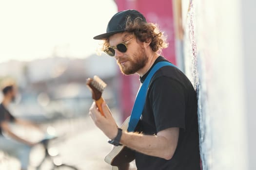 Smiling man hipster standing by the wall and playing guitar. Mid shot