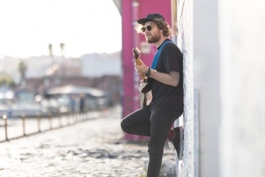 A man hipster standing by the wall and playing electric guitar. Mid shot