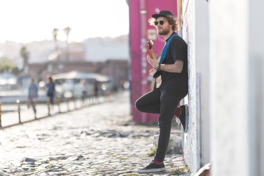 A man hipster standing by the wall and playing electric guitar on the street. Mid shot