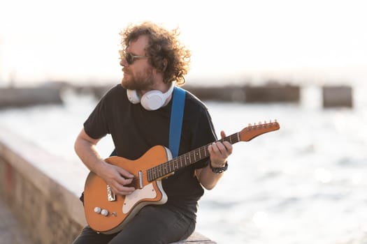 A man hipster wearing wireless headphones sitting at the waterfront and playing guitar. Mid shot