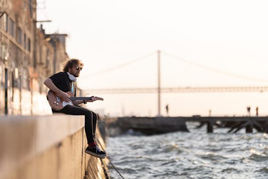 A man hipster sits on the embankment with his legs dangling to the water and plays the guitar. Mid shot