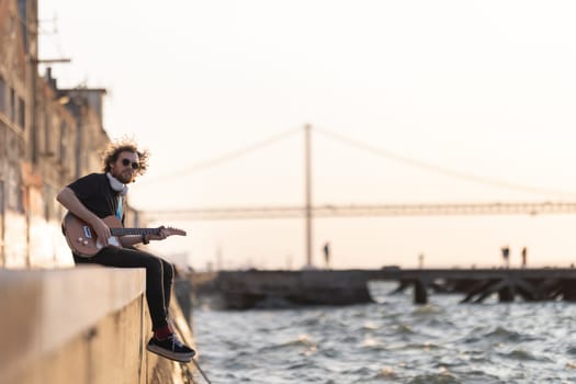 A man hipster sits on the waterfront with his legs dangling to the water and plays the guitar. Mid shot