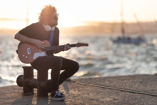 A man sits at the quay and playing guitar at early sunset. Mid shot