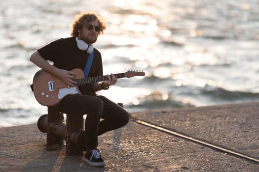 A man wearing wireless headphones sits at the quay and playing guitar at early sunset. Mid shot
