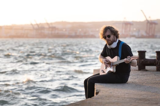 A man with curly hair wearing wireless headphones sits at the quay and playing guitar at early sunset. Mid shot