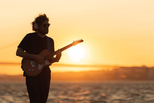 Romantic man stands at the waterfront and playing guitar at bright orange sunset. Mid shot