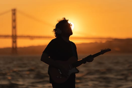 A shadow of romantic man standing at the waterfront and playing guitar at bright orange sunset. Mid shot
