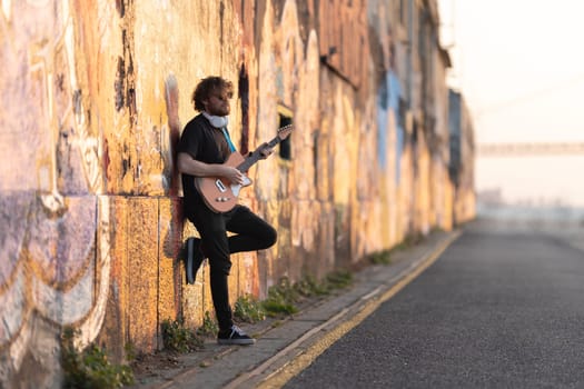 Romantic man hipster wearing sunglasses standing by the wall playing electric guitar at early sunset. Mid shot