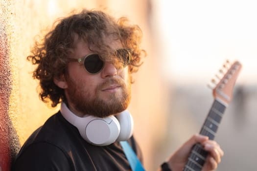 Romantic man with curly hair standing outdoors with a guitar at early sunset. Portrait
