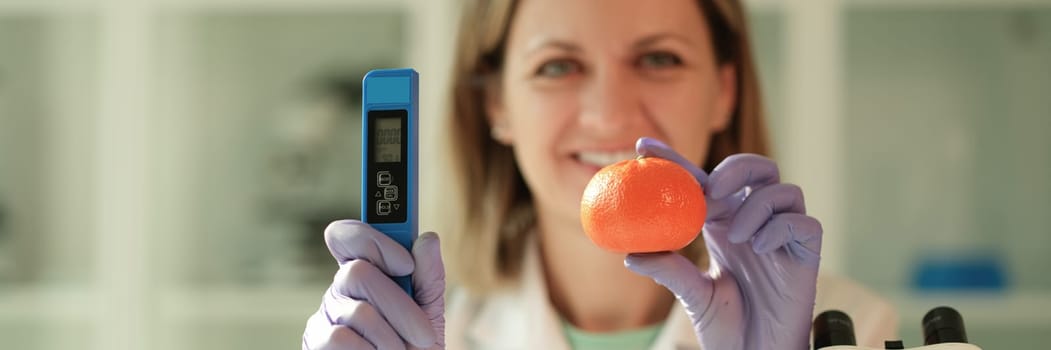 Scientist chemist holding in hands tangerine and device for measuring amount of nitrates in chemical laboratory closeup. Fruit and food quality control concept