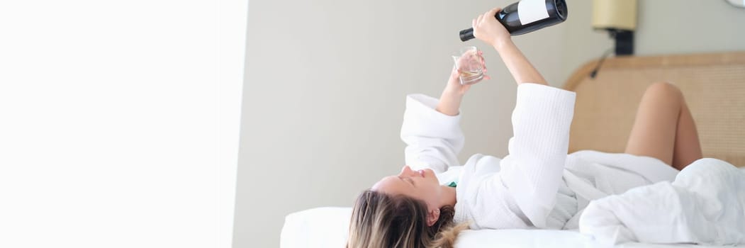 Woman in bathrobe lying in white bed and pouring champagne into glass. Rest and relaxation with alcohol in hotel concept