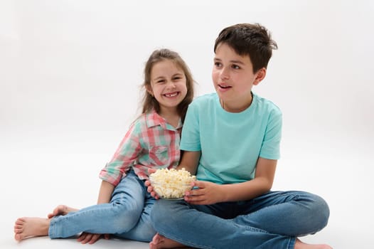 Mischievous adorable children, a teen boy and his younger sister sitting on white studio background, having fun together, eating popcorn and watching movie or cartoon. Kids and entertainment concept