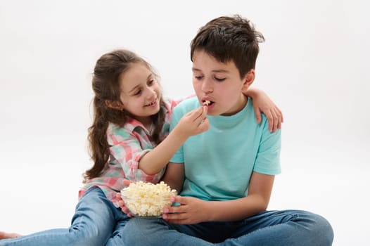 Lovely little girl hugging and feeding her older brother with tasty popcorn while watching movie or cartoons, isolated on white studio background. Kids. Entertainment. Leisure. Family relationships