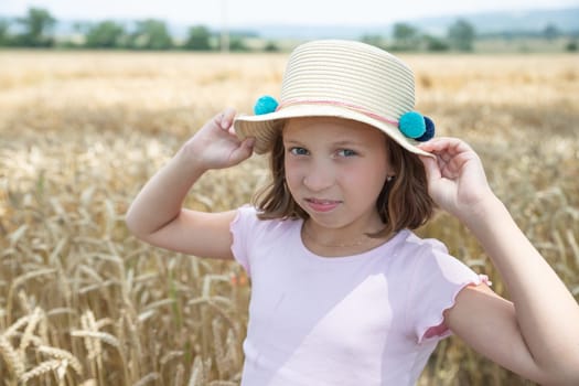 Adorable little blonde girl in a hat and a white linen dress, standing on a wheat field and touching the ears of wheat with her hands.