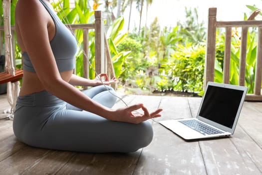 Unrecognizable woman in sports clothing following online meditation using laptop and headphones outdoors in tropical vacation resort. Spirituality and mental health concept.