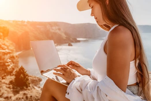 Digital nomad, Business woman working on laptop by the sea. Pretty lady typing on computer by the sea at sunset, makes a business transaction online from a distance. Freelance remote work on vacation