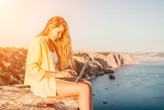 Successful business woman in yellow hat working on laptop by the sea. Pretty lady typing on computer at summer day outdoors. Freelance, travel and holidays concept.